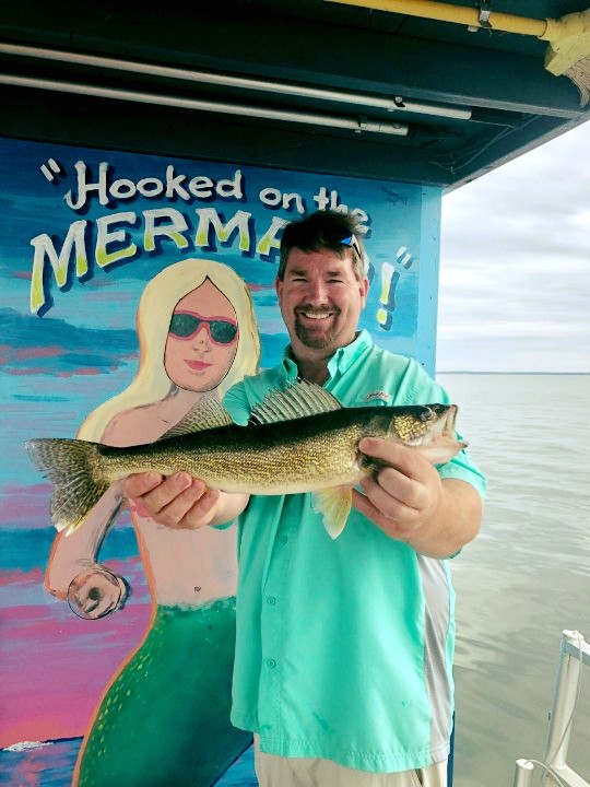 Man in greet t-shirt holding a fish in front of a sign saying hooked on the mermaid
